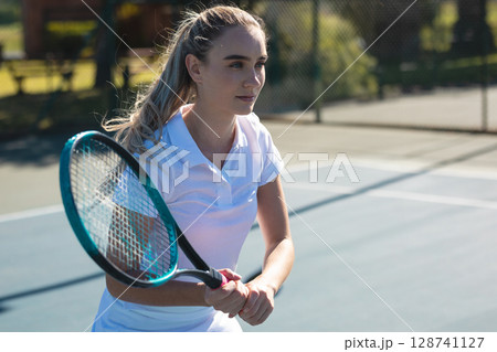 Young adult woman hitting backhand on outdoor tennis court, holding racket near chain-link fence 128741127