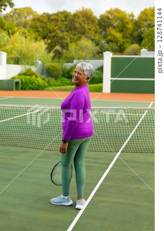 Senior woman practicing on outdoor tennis court, holding tennis racket and smiling by net and lines 128741144