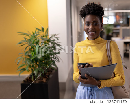 Young African American woman standing in office hallway, holding smartphone and folder, copy space Young African American woman standing in office hallway, holding smartphone and folder, copy space 128741170