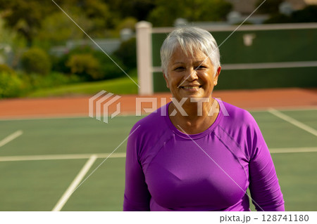 Senior woman standing and smiling on tennis court, with white lines and green fence rails Senior woman standing and smiling on tennis court, with white lines and green fence rails 128741180