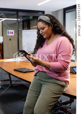 Woman leaning against wooden conference table in office, tapping tablet among laptops and documents 128741194