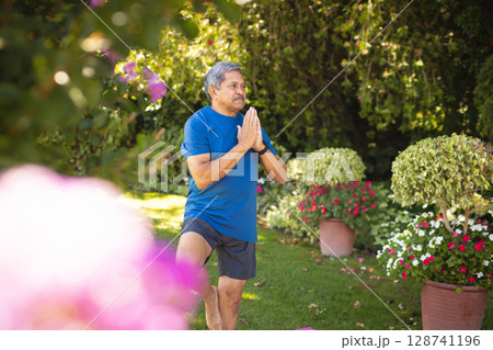 Senior man practicing tree pose on pink mat in sunny backyard, with potted flowers, copy space 128741196