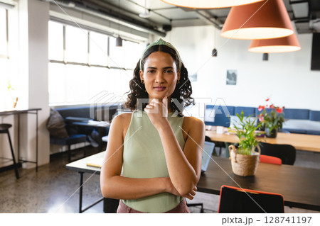 Woman standing thoughtfully in modern coworking office, surrounded by laptop, notebooks and plants 128741197