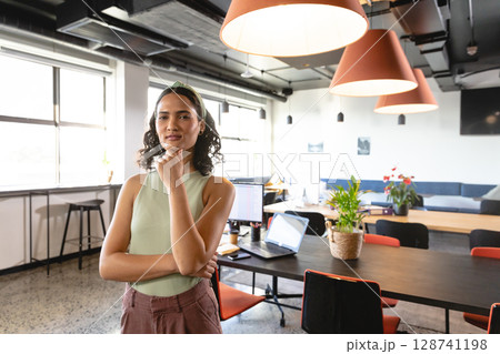 Professional woman standing confidently in modern open-plan office, with laptops and plants 128741198