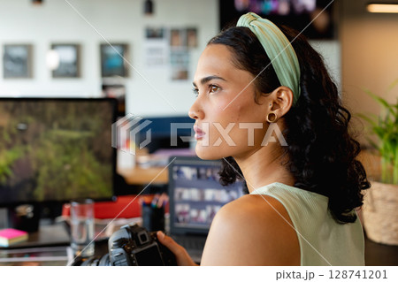 Female photographer holding DSLR camera at creative studio desk with monitors, supplies, copy space 128741201