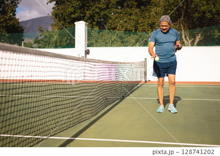 Senior man picking up yellow tennis balls near net on outdoor hard court, copy space 128741202