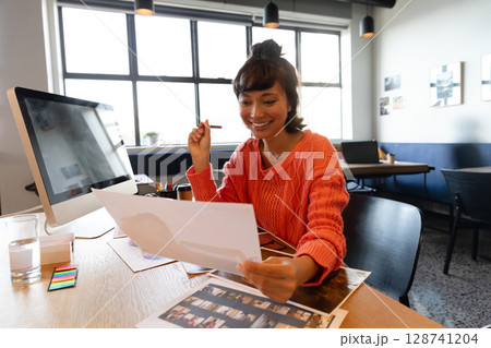 Young adult man sitting at wooden desk in open-plan office, holding sheet, smiling with smartphone 128741204