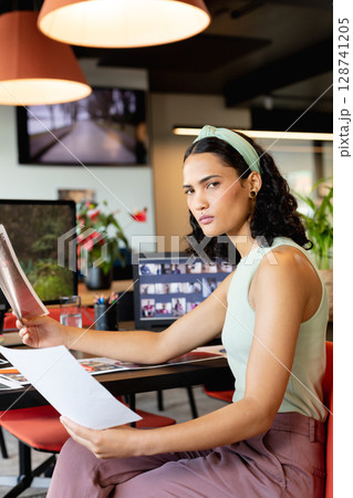 Woman reviewing printed sheets at open?plan office desk, surrounded by monitors and red chairs 128741205