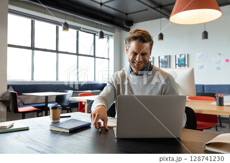 Man smiling while using silver laptop at open-plan office desk, holding coffee cup and pen 128741230