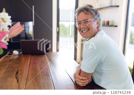Senior man sitting at wooden table in home, smiling at laptop with pink lilies, copy space 128741239