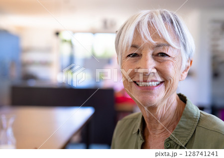 Senior woman smiling at camera in kitchen dining area, with wooden table and vase, copy space 128741241