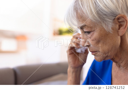 Senior woman wiping eye with white tissue in living room with gray sofa, copy space 128741252