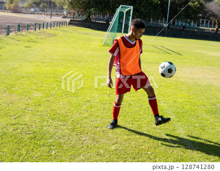 Male athlete juggling soccer ball on grass soccer pitch wearing red bib and cleats, copy space 128741280