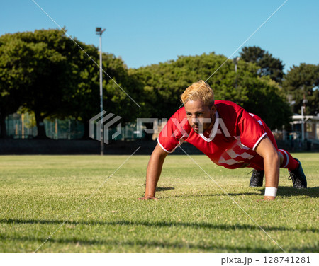 Man performing push-up on grass sports field, wearing red jersey white wristbands and black cleats Man performing push-up on grass sports field, wearing red jersey white wristbands and black cleats 128741281
