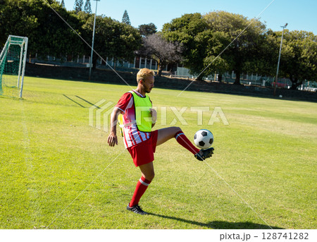 Male soccer player controlling soccer ball on grass soccer field, with goalpost and fluorescent bib 128741282