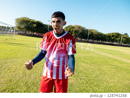 Young adult soccer player wearing striped jersey celebrating on grassy field, with white metal goal 128741288
