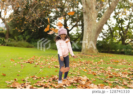 African American girl tossing autumn leaves in grassy park with pink hat and jacket, copy space 128741310