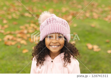African American girl smiling on grassy field in autumn, wearing pink pompom hat and pink jacket 128741311