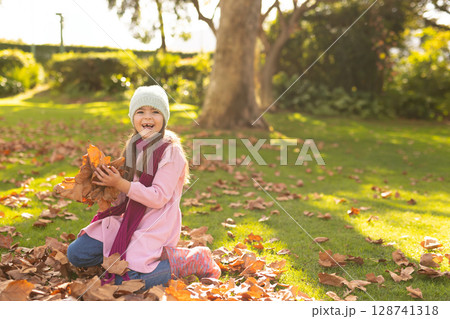 Girl kneeling on grassy park lawn, holding brown orange leaf pile with knit hat and boots Girl kneeling on grassy park lawn, holding brown orange leaf pile with knit hat and boots 128741318