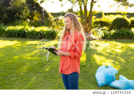 Woman standing and writing on black clipboard in sunny backyard with blue garbage bags, copy space Woman standing and writing on black clipboard in sunny backyard with blue garbage bags, copy space 128741328