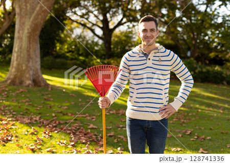 Man raking fallen leaves on sunlit grassy residential backyard lawn, holding red plastic leaf rake Man raking fallen leaves on sunlit grassy residential backyard lawn, holding red plastic leaf rake 128741336