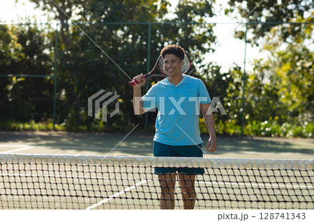 Mid-adult man standing behind tennis net on outdoor court, holding racket over court lines, smiling 128741343