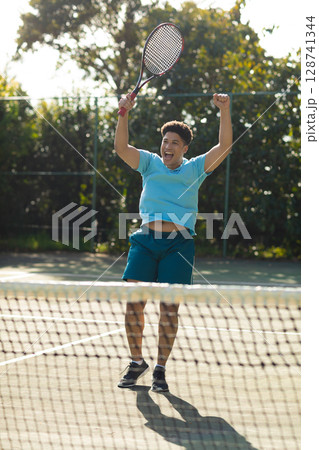 Man celebrating winning point on outdoor tennis court, jumping near net and holding racket 128741344