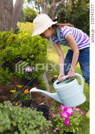 Child wearing straw hat watering flowers with light blue watering can in backyard garden bed 128741377