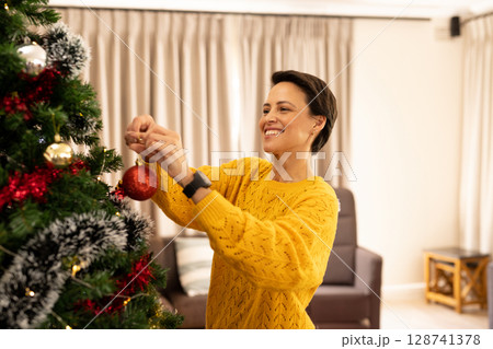 Woman hanging red bauble on decorated Christmas tree in living room, wearing smartwatch, smiling Woman hanging red bauble on decorated Christmas tree in living room, wearing smartwatch, smiling 128741378