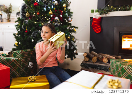 Female child sitting beside Christmas tree at home holding gold-wrapped present to ear, copy space Female child sitting beside Christmas tree at home holding gold-wrapped present to ear, copy space 128741379