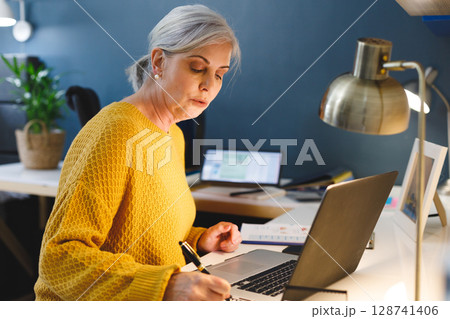 Woman writing in notebook in modern home office, using laptop with desk lamp and potted plant 128741406