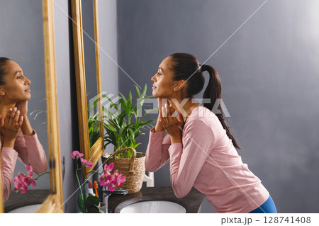 Young adult woman leaning on bathroom sink examining neck in mirror with orchids, copy space Young adult woman leaning on bathroom sink examining neck in mirror with orchids, copy space 128741408