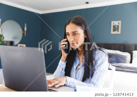 Woman talking on smartphone and typing on laptop at bedroom home office, with coffee mug Woman talking on smartphone and typing on laptop at bedroom home office, with coffee mug 128741413