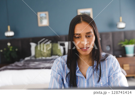 Young woman working on laptop at bedroom desk, surrounded by bed, plants and wall art Young woman working on laptop at bedroom desk, surrounded by bed, plants and wall art 128741414