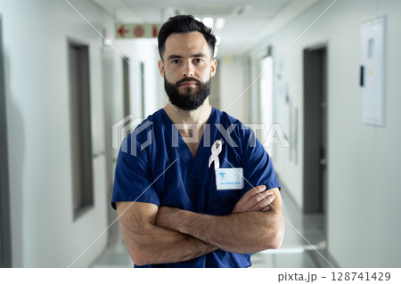 Young male doctor standing with arms crossed in hospital corridor, sporting badge and pink ribbon 128741429