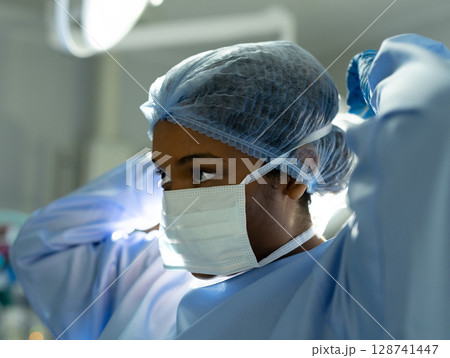 Female surgeon adjusting mask straps and surgical cap in operating room, with bright overhead light 128741447