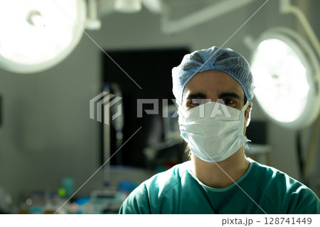 Male surgeon standing facing camera in operating room, wearing surgical mask and cap under lights 128741449