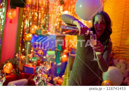 Woman pouring champagne into flute in party room, with balloons and gift boxes, copy space 128741453