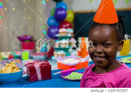 African American boy sitting at birthday party table, wearing party hat near cupcakes, copy space African American boy sitting at birthday party table, wearing party hat near cupcakes, copy space 128742207