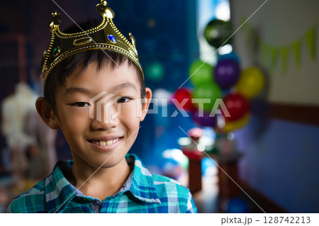 Chinese boy wearing gold crown and plaid shirt, smiling at home birthday celebration with balloons 128742213