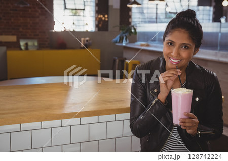 Indian man leaning against wood bar in cafe, sipping pink milkshake with whipped cream, copy space 128742224