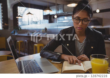 Indian woman reading open book at cafe table, tracing text with finger near laptop, copy space Indian woman reading open book at cafe table, tracing text with finger near laptop, copy space 128742228