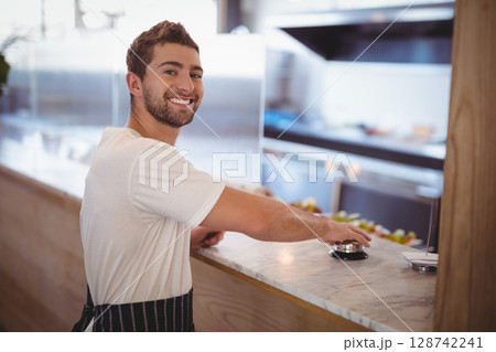 Young man smiling and pressing silver bell behind marble counter in cafe, with kitchen equipment 128742241