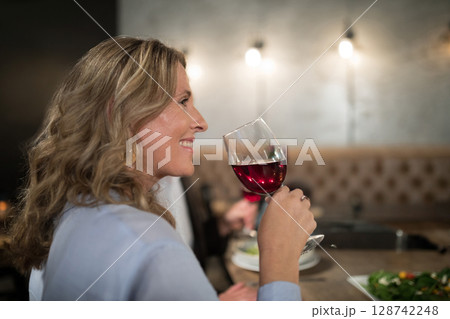 Senior woman holding stemmed wine glass in wine bar with wall sconces, enjoying salad beside bench Senior woman holding stemmed wine glass in wine bar with wall sconces, enjoying salad beside bench 128742248