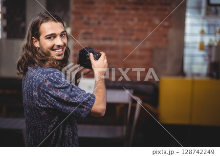 Young adult man smiling while holding DSLR camera in studio with open laptop and yellow cabinet Young adult man smiling while holding DSLR camera in studio with open laptop and yellow cabinet 128742249