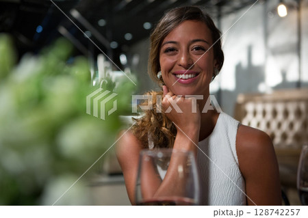 Woman resting chin on hand smiling at camera in restaurant with wine glass and green centerpiece 128742257