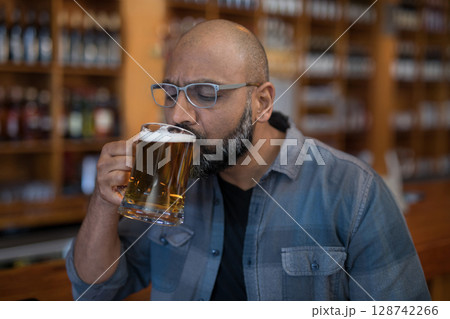 Indian man sipping beer from large glass mug at wooden bar counter, surrounded by bottled beers 128742266