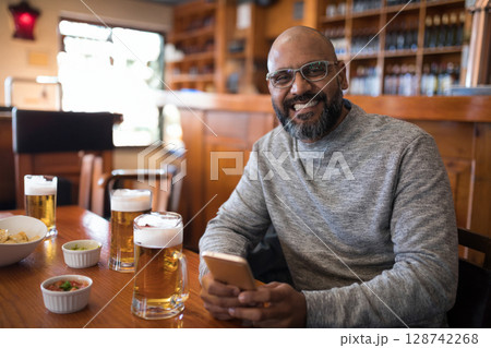 Indian man enjoying beer and chips with salsa at wooden pub table, smiling and using smartphone Indian man enjoying beer and chips with salsa at wooden pub table, smiling and using smartphone 128742268