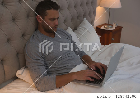 Mid-adult man typing on laptop in bedroom with tufted headboard, wooden nightstand lamp Mid-adult man typing on laptop in bedroom with tufted headboard, wooden nightstand lamp 128742300