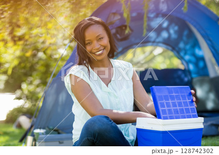 African American woman sitting on grass at campsite near blue dome tent, lifting cooler lid 128742302